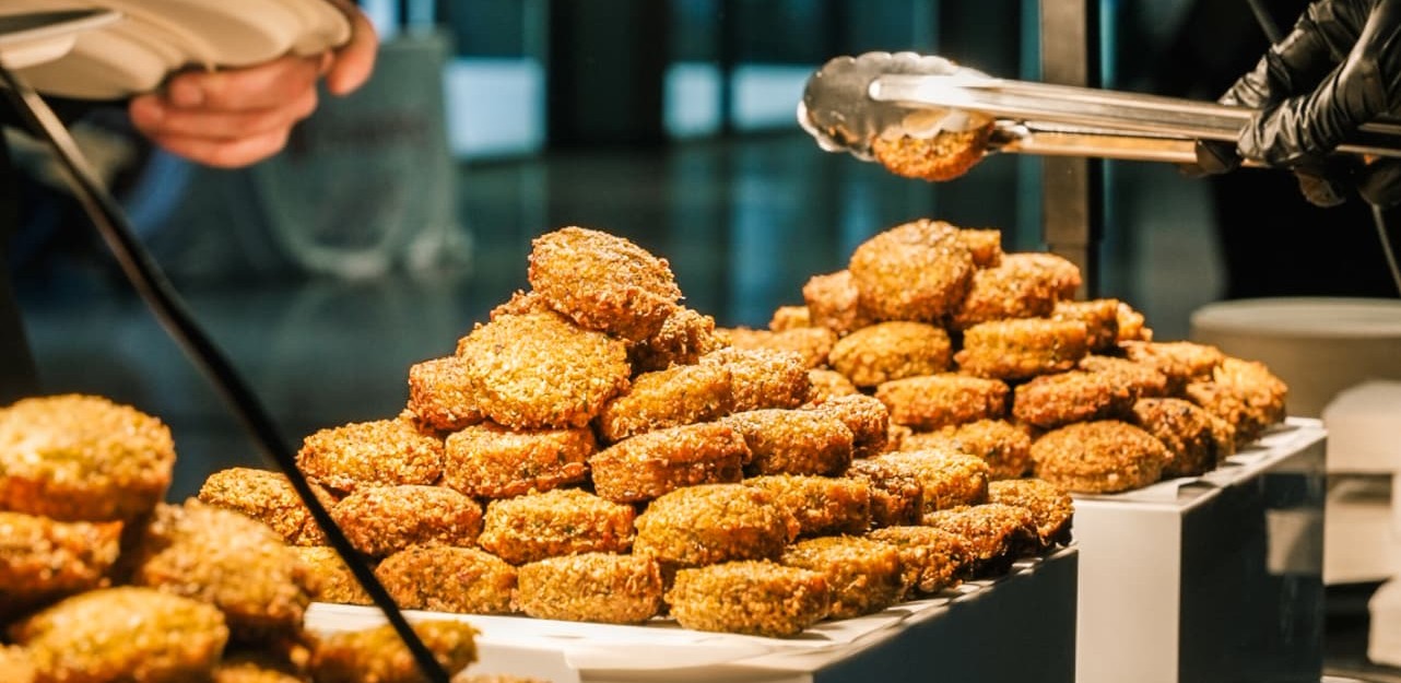 Golden-brown fritters stacked on white buffet risers under warm heat lamps, a server lifting one with tongs